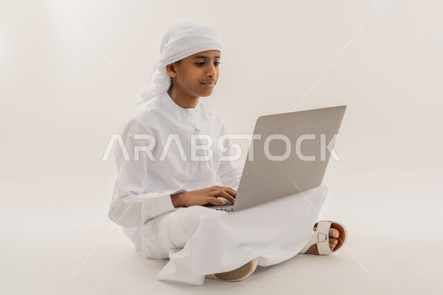 Using modern and advanced technology and techniques, distance learning and receiving lessons via the Internet, entertaining electronic entertainment activities, spending fun times, portrait of an Arab Gulf Emirati boy wearing a kandura and turban sitting on the floor and using a laptop, white background