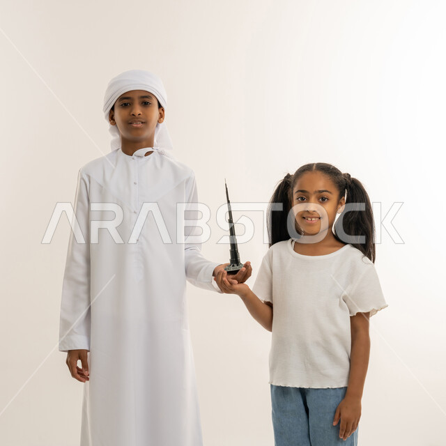 النظر للكاميرا بتعابيرالفرح، الأبراج الشاهقة وناطحات السحاب، معالم سياحية ترفيهية مشهورة بالإمارات، بورتريه مقرب لولد عربي خليجي اماراتي يرتدي الكندورة والعمامة يقف برفقة أخته ويقوموا بحمل مجسم ثلاثي الأبعاد لبرج خليفة الشهير، الفن الهندسي المعماري، خلفية بيضاء