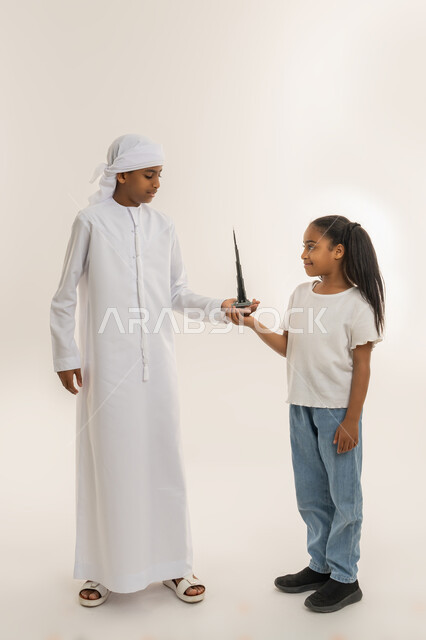 معالم سياحية ترفيهية مشهورة في الإمارات، الأبراج الشاهقة وناطحات السحاب، الفن الهندسي المعماري، بورتريه لولد عربي خليجي اماراتي يرتدي الكندورة والعمامة يقف برفقة أخته ويقوموا بحمل مجسم ثلاثي الأبعاد لبرج خليفة الشهير، صورة الجسم بالطول الكامل، خلفية بيضاء