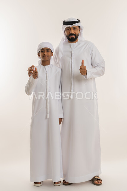 علامة الامتياز والجودة والنجاح، الشعور بالرضا والقبول، النظر للكاميرا بإيماءات السعادة والسرور، بورتريه لرجل عربي خليجي اماراتي يرتدي الكندورة والغترة يقف برفقة ابنه ويرفعوا اصبع الابهام للأعلى بتعابير الموافقة والاعجاب، صورة الجسم بالطول الكامل، خلفية بيضاء