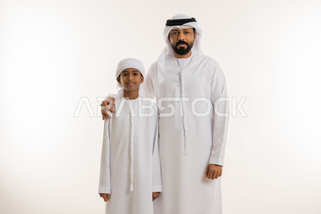 الشعور بالأمان مع الأب، مفهوم التربية القائمة على الحب والحنان، النظر الى الكاميرا بإيماءات السعادة والسرور، بورتريه مقرب لرجل عربي خليجي اماراتي يرتدي الكندورة والغترة البيضاء يقف برفقة ابنه ويضع يده على كتفه بتعابير المحبة والعطف، خلفية بيضاء