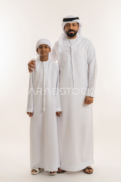 مفهوم التربية القائمة على الحب والحنان، الشعور بالأمان مع الأب، النظر الى الكاميرا بإيماءات السعادة والسرور، بورتريه لرجل عربي خليجي اماراتي يرتدي الكندورة والغترة البيضاء يقف برفقة ابنه ويضع يده على كتفه بتعابير المحبة، صورة الجسم بالطول الكامل، خلفية بيضاء