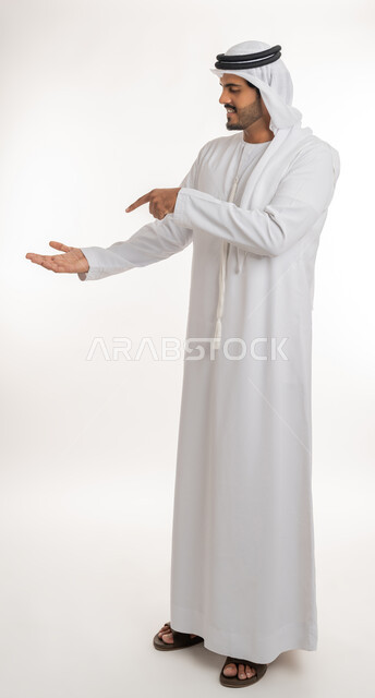 رفع وحمل عنصر معين, الاشارة بإصبع السبابة إلى شيء محدد, بورتريه لرجل عربي خليجي اماراتي يرتدي الكندورة والغترة البيضاء يقف باستقامة ويرفع يده ويشير بالإخرى بايماءات وجود شيء ما، الطول الكامل للجسم، خلفية بيضاء
