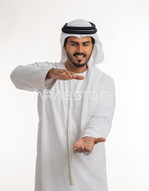 مفهوم الاناقة و الاهتمام بالمظهر الخارجي, إحتواء وإمساك عنصر ما،  بورتريه مقرب لرجل عربي خليجي اماراتي يرتدي الكندورة و العمامة يفتح يديه لعرض عناصر هولوجرامية ثلاثية الأبعاد، خلفية بيضاء