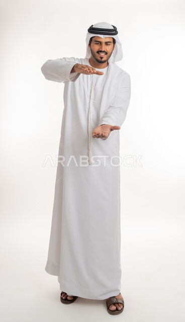 إحتواء وإمساك عنصر ما، مفهوم الاناقة و الاهتمام بالمظهر الخارجي, بورتريه لشاب عربي خليجي اماراتي يرتدي الكندورة و الغترة يفتح يديه لعرض عناصر هولوجرامية ثلاثية الأبعاد، صورة الجسم بالطول الكامل, خلفية بيضاء