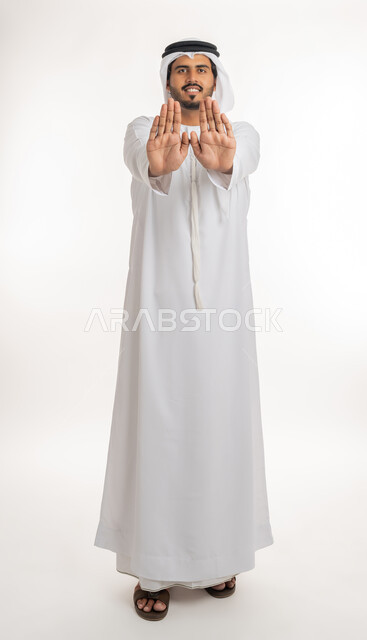 القلق والاحتياط والابتعاد عن شيء ما, حماية النفس من ضرر خارجي, بورتريه لرجل  عربي خليجي اماراتي يرتدي الكندورة والغترة البيضاء يرفع يديه للاعلى بإيماءات التوقف وعدم الاقتراب، مفهوم الحذر وترك مسافة، خلفية بيضاء