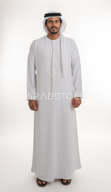 الوقوف باستقامه والنظر الى الكاميرا بتعابير الثقه بالنفس، مفهوم الاناقه والاهتمام بالمظهر الخارجي، بورتريه لرجل عربي خليجي اماراتي يرتدي الكندوره والغتره البيضاء الاماراتيه يقف بايماءات الرجولة، خلفية بيضاء