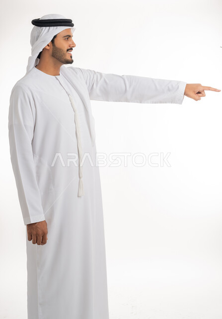 Indicating the location of an item, pointing and explaining something, specifying a specific location, portrait of an Arab Gulf Emirati man wearing a white kandura and ghutra pointing with his index finger to something, white background