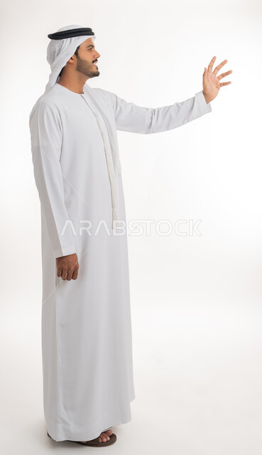 الإشارة إلى عنصر ما، بورتريه لرجل عربي خليجي اماراتي يرتدي الكندورة و الغترة البيضاء يمد يده للأمام يحاول التقاط عناصر هولوجرامية ثلاثية الأبعاد، محاولة الإحاطة والإمساك بشيء ما، الجسم بالطول الكامل، خلفية بيضاء