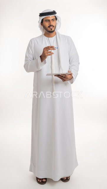 Blank green chroma key display on technical devices, using tablet to complete work, using modern and advanced technology for learning, portrait of an Arab Gulf Emirati man wearing kandura and ghutra holding a tablet in his hand, full body, white background
