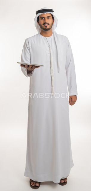 Blank green chroma key display on technical devices, using tablet to complete work, using modern and advanced technology for learning, portrait of an Arab Gulf Emirati man wearing kandura and ghutra holding a tablet in his hand, full body, white background