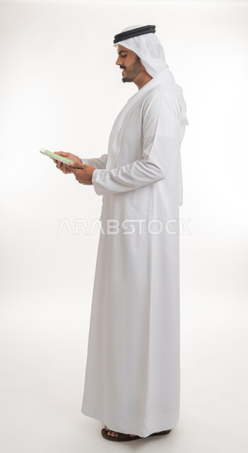استعمال الجهاز اللوحي في إنجاز العمل، عرض شاشة كروما خضراء فارغة على الأجهزة التقنية، استعمال التكنولوجيا الحديثة والمتطورة للتعلم، بورتريه لرجل عربي خليجي اماراتي يرتدي الكندورة والغترة يمسك بيده تابلت، الجسم بالطول الكامل، خلفية بيضاء