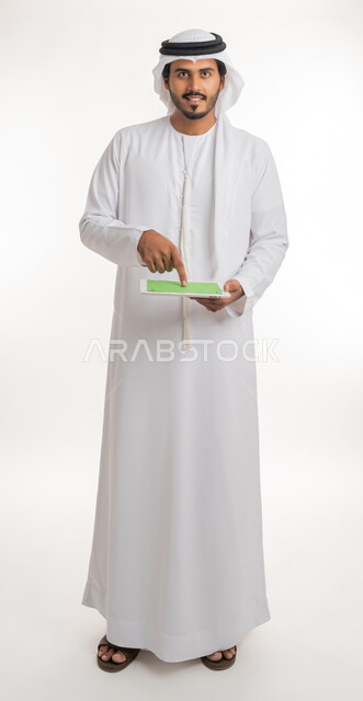 Blank green chroma key display on technical devices, using tablet to complete work, using modern and advanced technology for learning, portrait of an Arab Gulf Emirati man wearing kandura and ghutra holding a tablet in his hand, full body, white background