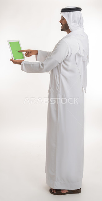 استعمال الجهاز اللوحي في إنجاز العمل، عرض شاشة كروما خضراء فارغة على الأجهزة التقنية، استعمال التكنولوجيا الحديثة والمتطورة للتعلم، بورتريه لرجل عربي خليجي اماراتي يرتدي الكندورة والغترة يمسك بيده تابلت، الجسم بالطول الكامل، خلفية بيضاء