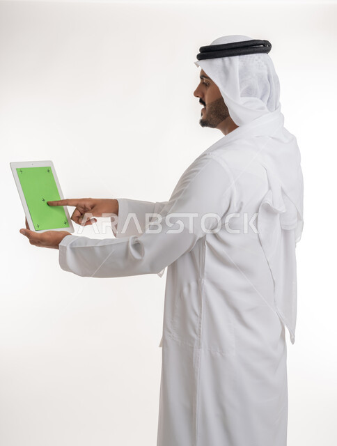 Using modern and advanced technology for learning, displaying a blank green chroma key screen on technical devices, using a tablet to complete work, close-up portrait of an Arab Gulf Emirati man wearing a kandura and ghutra holding a tablet in his hand, white background