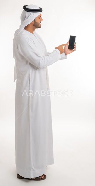 تصفح مواقع التواصل الاجتماعي عبر الانترنت، إجراء مكالمات فيديو عبر الجوال، تطور التكنولوجيا والتقنيات الحديثة، بورتريه من الجانب لرجل اماراتي عربي خليجي يرتدي الكندورة والغترة يقوم باستخدام الهاتف المحمول، صورة الجسم بالطول الكامل, خلفية بيضاء