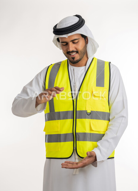 Working in the engineering sector, concept of engineering and architectural construction, containing and holding an element, portrait of an Arab Gulf Emirati engineer wearing a white kandura and ghutra with a protective jacket, opening his hands to display 3D holographic elements, white background