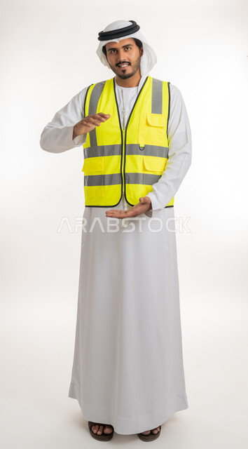 العمل في القطاع الهندسي، مفهوم الهندسة والانشاءات المعمارية، إحتواء وإمساك عنصر ما، بورتريه لمهندس عربي خليجي اماراتي يرتدي الكندورة والغترة البيضاء مع سترة الحماية يفتح يديه لعرض عناصرهولوجرامية ثلاثية الأبعاد، خلفية بيضاء