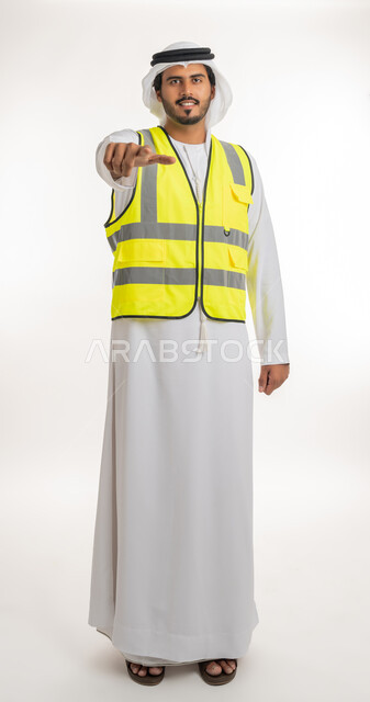 إشارات تدل على هذا المكان(هنا)، مفهوم الهندسة والانشاءات المعمارية، تحديد موقع معين، العمل في القطاع الهندسي، بورتريه لمهندس عربي خليجي اماراتي يرتدي الكندورة والغترة البيضاء مع سترة الحماية يرفع اصابعه ويشير الى شيء ما، خلفية بيضاء