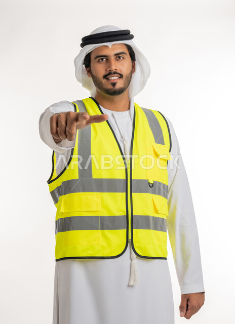 إشارات تدل على هذا المكان(هنا)، مفهوم الهندسة والانشاءات المعمارية، تحديد موقع معين، العمل في القطاع الهندسي، بورتريه لمهندس عربي خليجي اماراتي يرتدي الكندورة والغترة البيضاء مع سترة الحماية يرفع اصابعه ويشير الى شيء ما، خلفية بيضاء