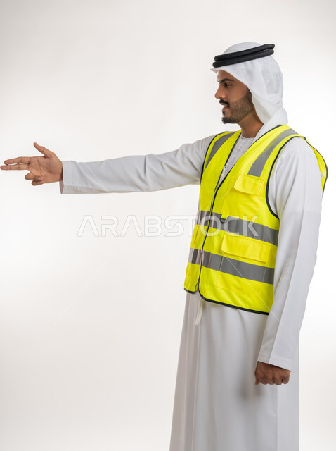 إشارات تدل على هذا المكان(هنا)، مفهوم الهندسة والانشاءات المعمارية، تحديد موقع معين، العمل في القطاع الهندسي، بورتريه لمهندس عربي خليجي اماراتي يرتدي الكندورة والغترة البيضاء مع سترة الحماية يرفع اصابعه ويشير الى شيء ما، خلفية بيضاء