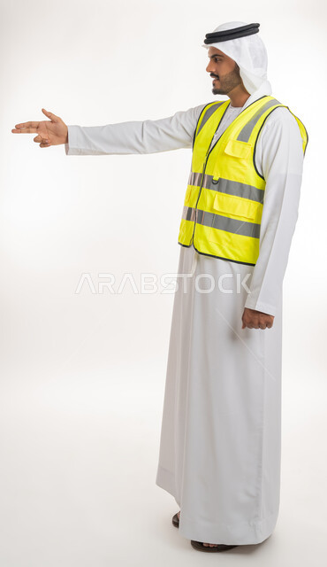 إشارات تدل على هذا المكان(هنا)، مفهوم الهندسة والانشاءات المعمارية، تحديد موقع معين، العمل في القطاع الهندسي، بورتريه لمهندس عربي خليجي اماراتي يرتدي الكندورة والغترة البيضاء مع سترة الحماية يرفع اصابعه ويشير الى شيء ما، خلفية بيضاء