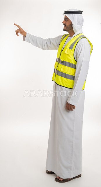 إشارات تدل على هذا المكان(هنا)، مفهوم الهندسة والانشاءات المعمارية، تحديد موقع معين، العمل في القطاع الهندسي، بورتريه لمهندس عربي خليجي اماراتي يرتدي الكندورة والغترة البيضاء مع سترة الحماية يرفع اصابعه ويشير الى شيء ما، خلفية بيضاء