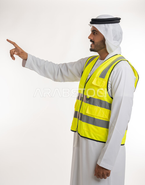 إشارات تدل على هذا المكان(هنا)، مفهوم الهندسة والانشاءات المعمارية، تحديد موقع معين، العمل في القطاع الهندسي، بورتريه لمهندس عربي خليجي اماراتي يرتدي الكندورة والغترة البيضاء مع سترة الحماية يرفع اصابعه ويشير الى شيء ما، خلفية بيضاء