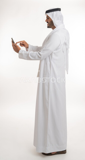 تصفح مواقع التواصل الاجتماعي عبر الانترنت، تطور التكنولوجيا والتقنيات الحديثة، بورتريه من الجانب لرجل اماراتي عربي خليجي يرتدي الكندورة والغترة يقوم باستخدام الهاتف المحمول، إجراء مكالمات فيديو عبر الجوال، صورة الجسم بالطول الكامل, خلفية بيضاء
