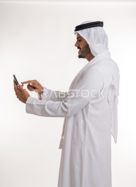 The development of technology and modern techniques, browsing social networking sites online, a portrait from the side of an Emirati, Arab, Gulf man wearing a kandora and ghutra, using a mobile phone, making video calls via mobile, full-length body photo, white background