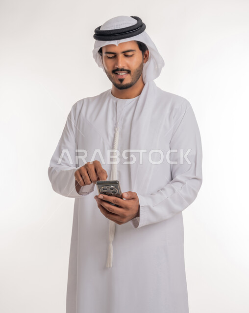 Making video calls via mobile phone, browsing social networking sites via the Internet, the development of technology and modern techniques, a portrait from the side of an Emirati Arab Gulf man wearing a kandora and ghutra, using a mobile phone, white background