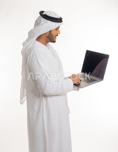 متابعة الأفلام والمسلسلات، تصفح مواقع التواصل الإجتماعي، الاجتهاد وانجاز الاعمال المطلوبة، بورتريه جانبي لرجل عربي خليجي اماراتي يرتدي الكندورة والغترة البيضاء يقف باستقامة يمسك بيده جهاز الحاسوب المحمول، استخدام التقنيات الحديثه والعمل عن بعد، خلفية بيضاء
