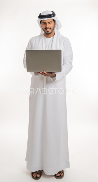 Watching movies and series, browsing social media, working hard and completing the required tasks, side portrait of an Arab Gulf Emirati man wearing a white kandura and ghutra, standing straight holding a laptop in his hand, using modern technologies and working remotely, white background