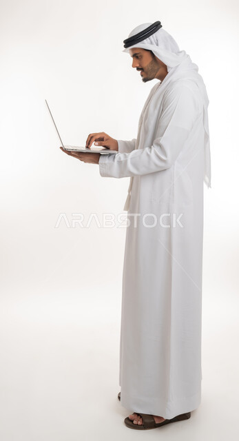Watching movies and series, browsing social media, working hard and completing the required tasks, side portrait of an Arab Gulf Emirati man wearing a white kandura and ghutra, standing straight holding a laptop in his hand, using modern technologies and working remotely, white background