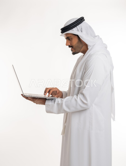 Watching movies and series, browsing social media, working hard and completing the required tasks, side portrait of an Arab Gulf Emirati man wearing a white kandura and ghutra, standing straight holding a laptop in his hand, using modern technologies and working remotely, white background