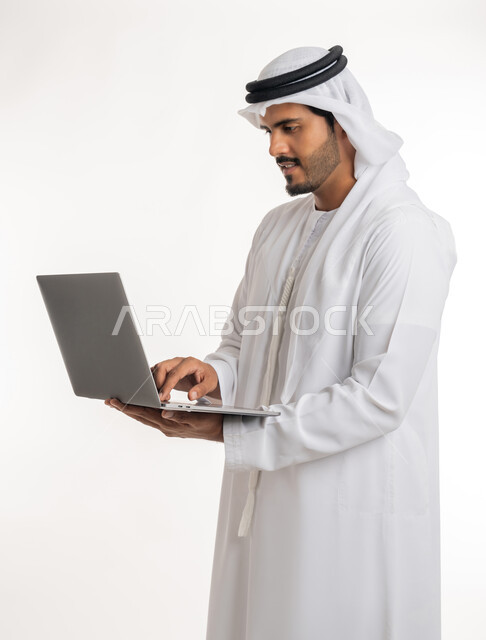 متابعة الأفلام والمسلسلات، تصفح مواقع التواصل الإجتماعي، الاجتهاد وانجاز الاعمال المطلوبة، بورتريه جانبي لرجل عربي خليجي اماراتي يرتدي الكندورة والغترة البيضاء يقف باستقامة يمسك بيده جهاز الحاسوب المحمول، استخدام التقنيات الحديثه والعمل عن بعد، خلفية بيضاء