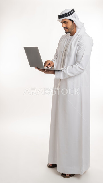 Watching movies and series, browsing social media, working hard and completing the required tasks, side portrait of an Arab Gulf Emirati man wearing a white kandura and ghutra, standing straight holding a laptop in his hand, using modern technologies and working remotely, white background