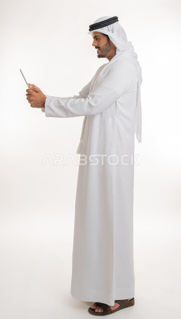 The concept of integrating work with technology and technology, completing work tasks using a tablet device, Emirati companies and jobs, achieving company goals, a side portrait of a Gulf Arab Emirati man wearing a kandora with a ghutra, full-length body photo, white background