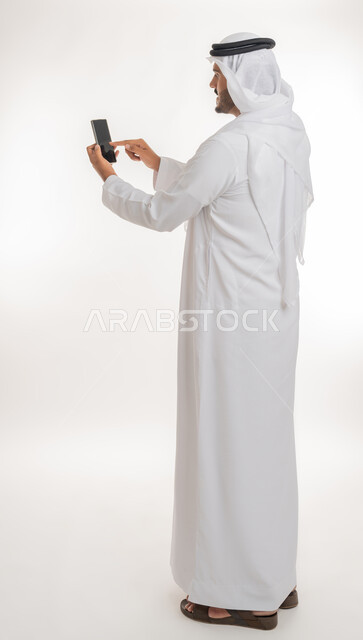 تطور التكنولوجيا والتقنيات الحديثة، تصفح مواقع التواصل الاجتماعي عبر الانترنت،  بورتريه من الخلف لرجل اماراتي عربي خليجي يرتدي الكندورة والغترة يقوم باستخدام الهاتف المحمول، إجراء مكالمات فيديو عبر الجوال، صورة الجسم بالطول الكامل, خلفية بيضاء