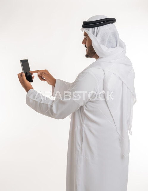 تصفح مواقع التواصل الاجتماعي عبر الانترنت، تطور التكنولوجيا والتقنيات الحديثة، بورتريه من الخلف لرجل اماراتي عربي خليجي يرتدي الكندورة والغترة يقوم باستخدام الهاتف المحمول، إجراء مكالمات فيديو عبر الجوال، خلفية بيضاء
