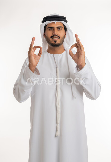 Joy, looks of enthusiasm and a sense of satisfaction, approval, acceptance and advice, expression of quality and mastery, portrait of a smiling Arab Gulf Emirati young man wearing the Emirati kandora and ghutra, raising his hands with gestures of quality, excellence and distinction, white background.