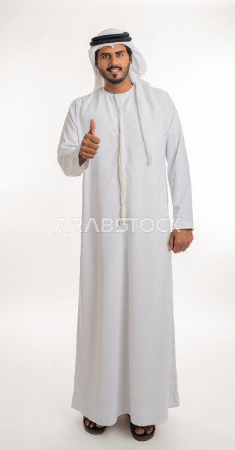 الرضا بالنتيجه الإمتياز والتميز، إيماءات وجه ويدين تدل على الموافقه والقبول، بورتريه مقرب لرجل عربي خليجي اماراتي يرتدي الثوب التقليدي الإماراتي والغترة البيضاء، يرفع أصبع الإبهام الى أعلى، الإعجاب والرضى والنجاح, الجسم بالطول الكامل، خلفية بيضاء