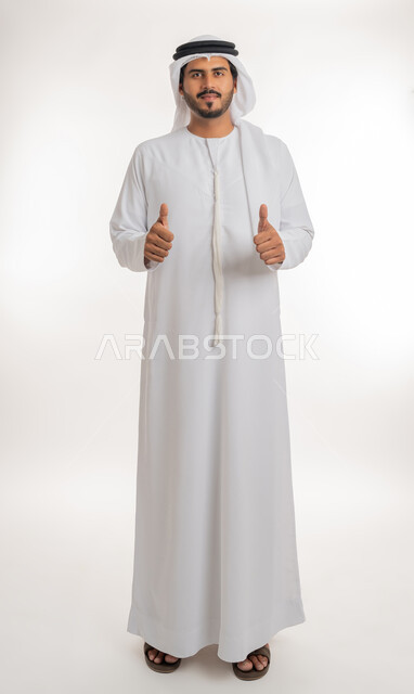 الرضا بالنتيجه الإمتياز والتميز، إيماءات وجه ويدين تدل على الموافقه والقبول، بورتريه مقرب لرجل عربي خليجي اماراتي يرتدي الثوب التقليدي الإماراتي والغترة البيضاء، يرفع أصبع الإبهام الى أعلى، الإعجاب والرضى والنجاح, الجسم بالطول الكامل، خلفية بيضاء