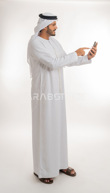 إجراء مكالمات فيديو عبر الجوال، تطور التكنولوجيا والتقنيات الحديثة، تصفح مواقع التواصل الاجتماعي عبر الانترنت،  بورتريه من الجانب لرجل اماراتي عربي خليجي يرتدي الكندورة والغترة يقوم باستخدام الهاتف المحمول، صورة الجسم بالطول الكامل, خلفية بيضاء