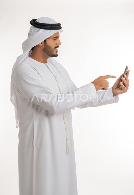 Browsing social networking sites online, the development of technology and modern techniques, a portrait of an Emirati, Arab, Gulf man wearing a kandora and ghutra, using a mobile phone, making video calls via mobile, white background