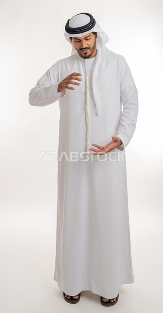تعابير السرور والسعادة, مفهوم الاناقة و الاهتمام بالمظهر الخارجي, إحتواء وإمساك عنصر ما، بورتريه  لرجل عربي خليجي اماراتي يرتدي الكندورة والغترة يفتح يديه لعرض عناصر هولوجرامية ثلاثية الأبعاد، صورة الجسم بالطول الكامل, خلفية بيضاء