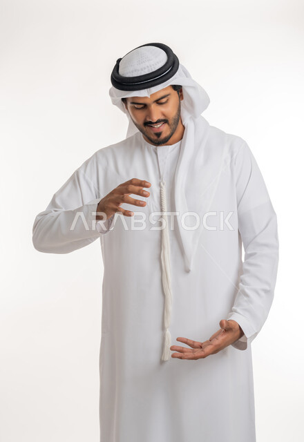 مفهوم الاناقة و الاهتمام بالمظهر الخارجي, إحتواء وإمساك عنصر ما،  بورتريه مقرب لرجل عربي خليجي اماراتي يرتدي الكندورة و العمامة يفتح يديه لعرض عناصر هولوجرامية ثلاثية الأبعاد، خلفية بيضاء