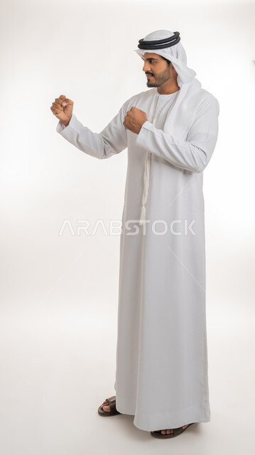 ممارسة رياضة الدفاع عن النفس، تعزيز صحة العظام والمفاصل، إتقان فنون الملاكمة، زيادة قدرة العضلات على التحمل، بورتريه من الجانب لرجل عربي خليجي اماراتي يرتدي الكندورة والغترة يرفع قبضات يديه بإيماءات القوة، صورة الجسم بالطول الكامل, خلفية بيضاء