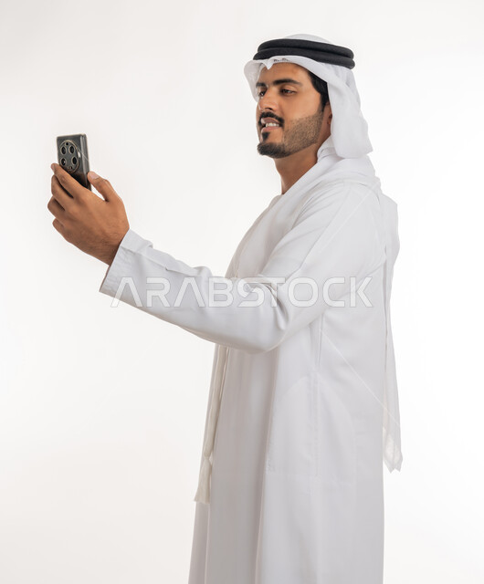 Browsing social networking sites online, the development of technology and modern techniques, a portrait of an Emirati, Arab, Gulf man wearing a kandora and ghutra, using a mobile phone, making video calls via mobile, white background