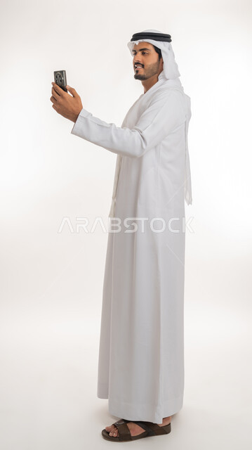 إجراء مكالمات فيديو عبر الجوال، تطور التكنولوجيا والتقنيات الحديثة، تصفح مواقع التواصل الاجتماعي عبر الانترنت،  بورتريه لرجل اماراتي عربي خليجي يرتدي الكندورة والغترة يقوم باستخدام الهاتف المحمول، صورة الجسم بالطول الكامل, خلفية بيضاء